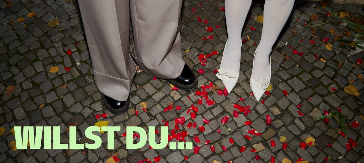 Schuhe von einem Ehepaar, das auf Rosenblättern steht, darunter der Schriftzug "Willst du..."